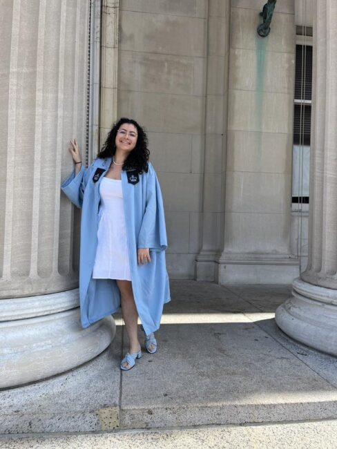 Female student in graduation gown stands against column