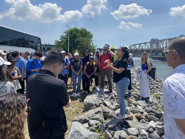 Group stands along a shoreline and listens to a woman speak