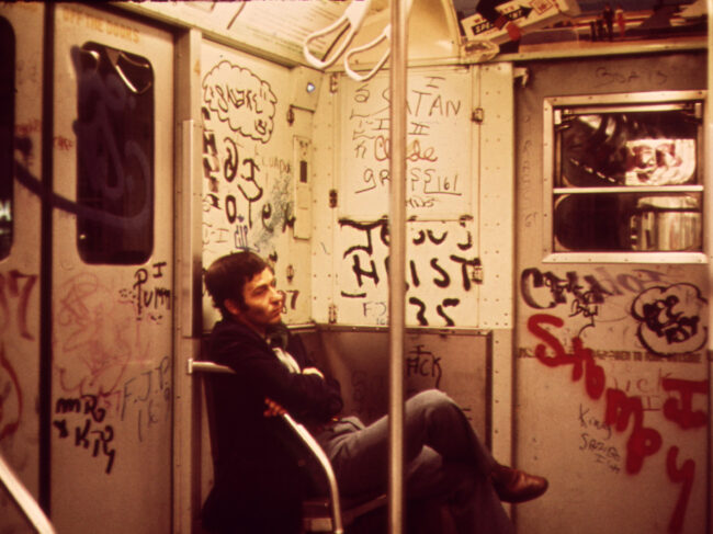 A man sitting in a subway car with heavy graffiti, 1973