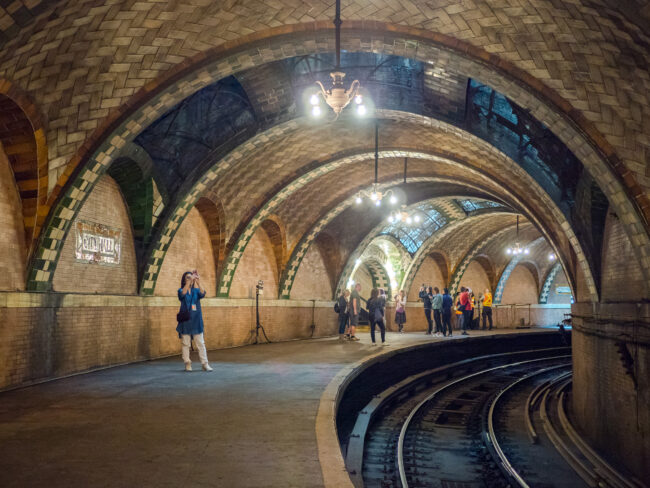 A grand subway tunnel with arches and chandeliers next to a curved section of track.