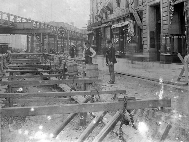 An old black and white photo with two men standing next to construction beams in the middle of a dug up street.