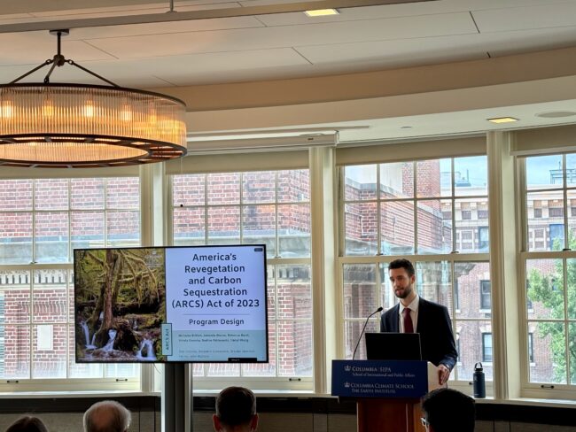 Man in suit and tie presenting at podium with a slideshow