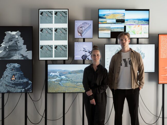Two people stand in front of screens with wires against a wall