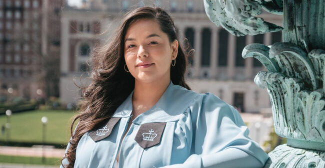 A student in a blue Columbia graduation gown posing on campus